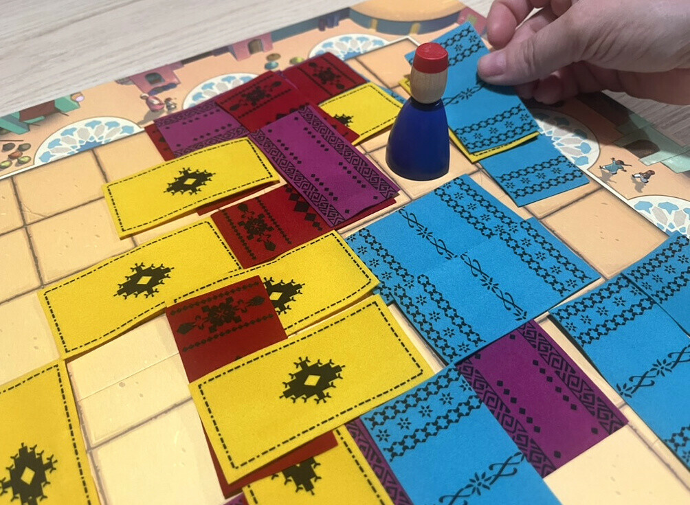 Une main pose un tapis bleu près d’Assam sur un plateau déjà couvert de tapis.